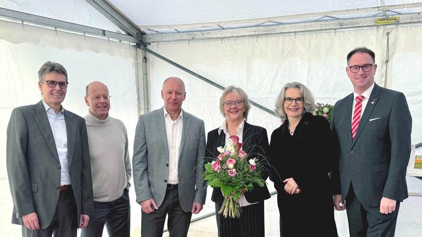 Eine Gruppe von sechs Personen steht nebeneinander, vier Männer und zwei Frauen. Eine der Frauen trägt einen Blumenstrauß. Die Gruppe steht in der Ecke eines Pagodenzelts.