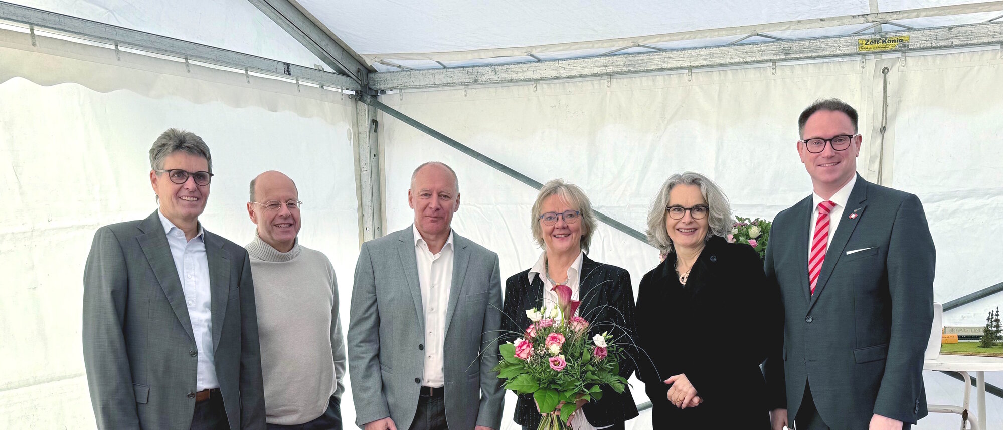 Eine Gruppe von sechs Personen steht nebeneinander, vier Männer und zwei Frauen. Eine der Frauen trägt einen Blumenstrauß. Die Gruppe steht in der Ecke eines Pagodenzelts.